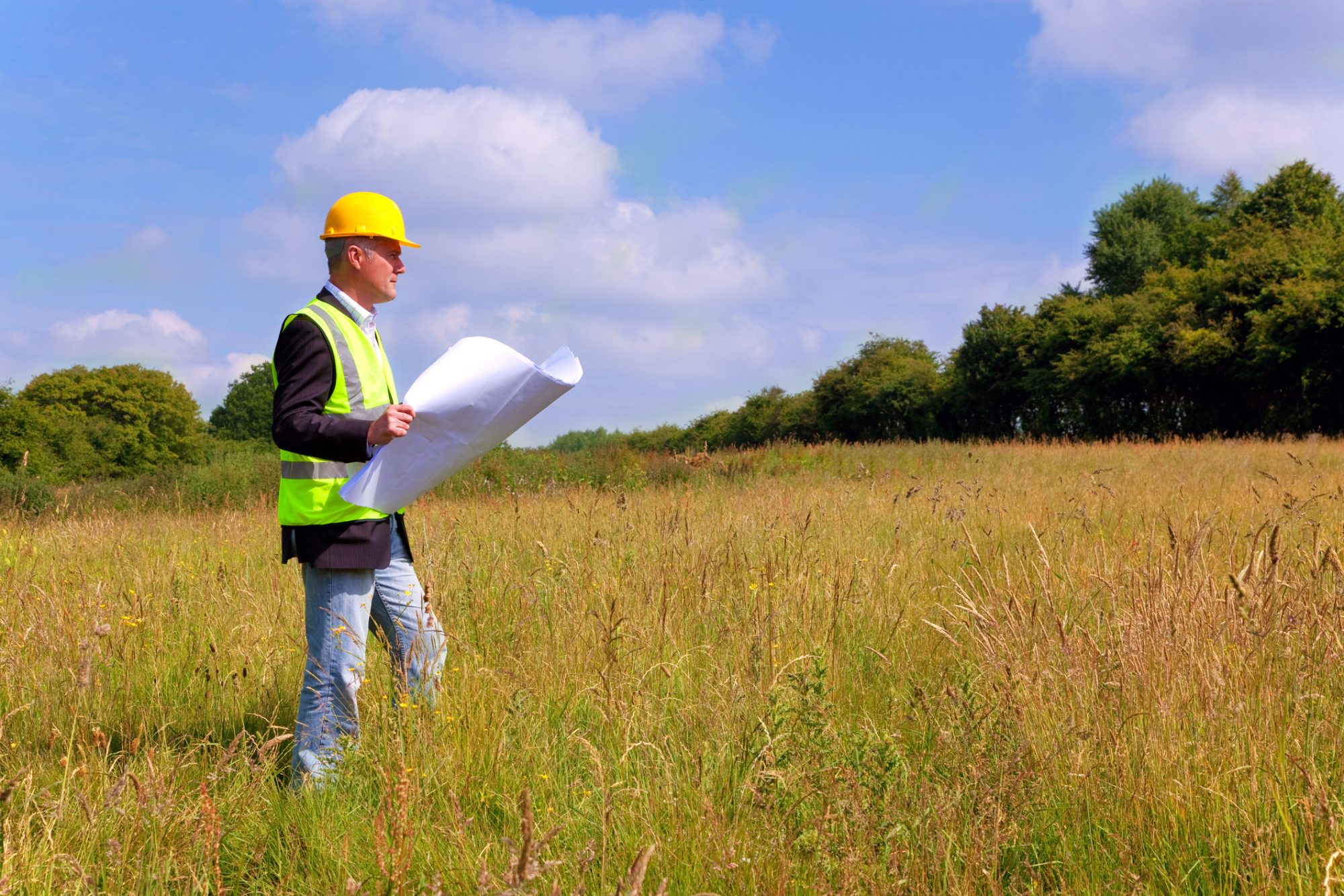 Architect wearing site safety gear and holding plans surveying a new building plot